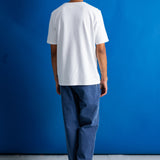 A man viewed from behind wearing a plain white short-sleeve t-shirt, loose blue jeans, and black shoes, standing against a solid blue studio backdrop.