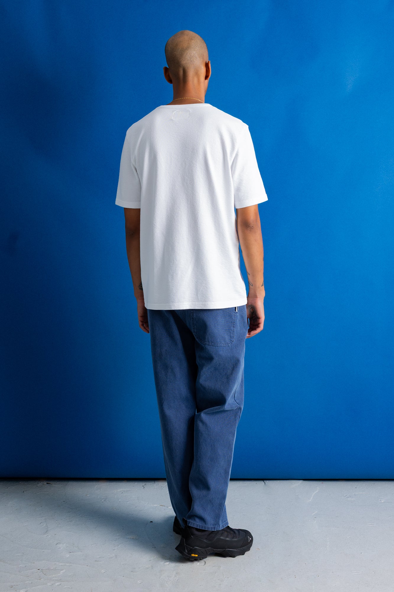 A man viewed from behind wearing a plain white short-sleeve t-shirt, loose blue jeans, and black shoes, standing against a solid blue studio backdrop.