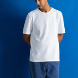 A man wearing a plain white short-sleeve crewneck t-shirt and blue jeans, standing against a solid blue studio background.