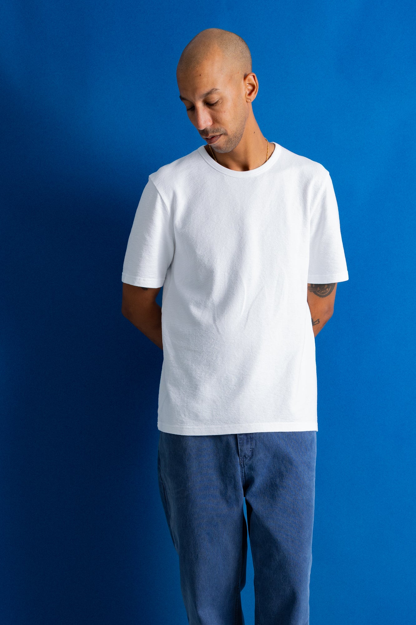 A man wearing a plain white short-sleeve crewneck t-shirt and blue jeans, standing against a solid blue studio background.
