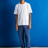A man stands against a bright blue studio backdrop wearing a plain white short-sleeve crewneck t-shirt, blue jeans, and black shoes.