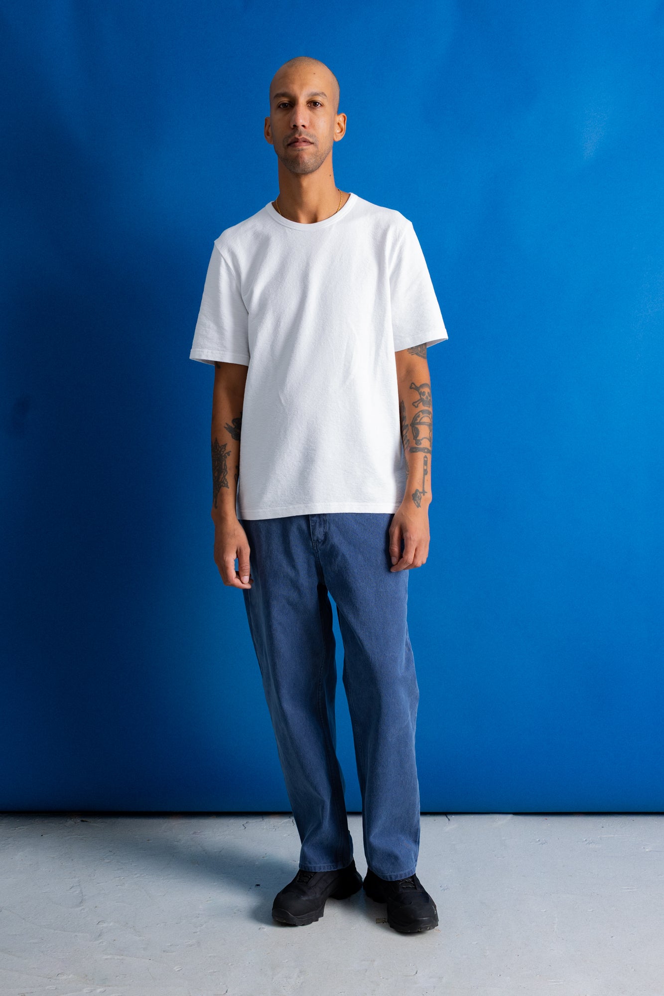 A man stands against a bright blue studio backdrop wearing a plain white short-sleeve crewneck t-shirt, blue jeans, and black shoes.