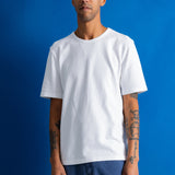 A man wearing a plain white crew-neck t-shirt and blue pants, standing against a solid blue background.