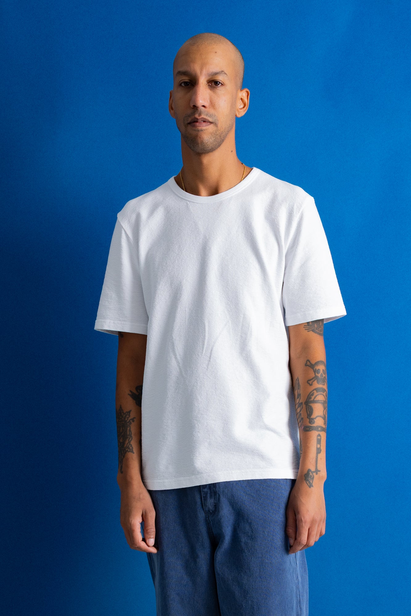 A man wearing a plain white crew-neck t-shirt and blue pants, standing against a solid blue background.