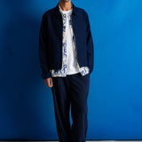 A man stands against a bright blue studio backdrop wearing a dark navy jacket over a white shirt with a blue patterned print, dark navy trousers, and black shoes.