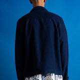 A man viewed from behind wearing a dark navy blue jacket over a white shirt with a blue abstract print, set against a solid blue studio background.