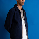 A man wearing a dark navy blue zip-up jacket over a patterned button-down shirt and white t-shirt, paired with beige pants, standing against a solid blue background.