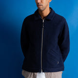 A man wearing a dark navy blue zip-up fleece jacket over a patterned shirt and beige pants, standing against a solid blue background.