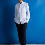 A man wearing a light blue long-sleeve button-down shirt, dark trousers, and black shoes, standing against a vibrant blue studio backdrop.