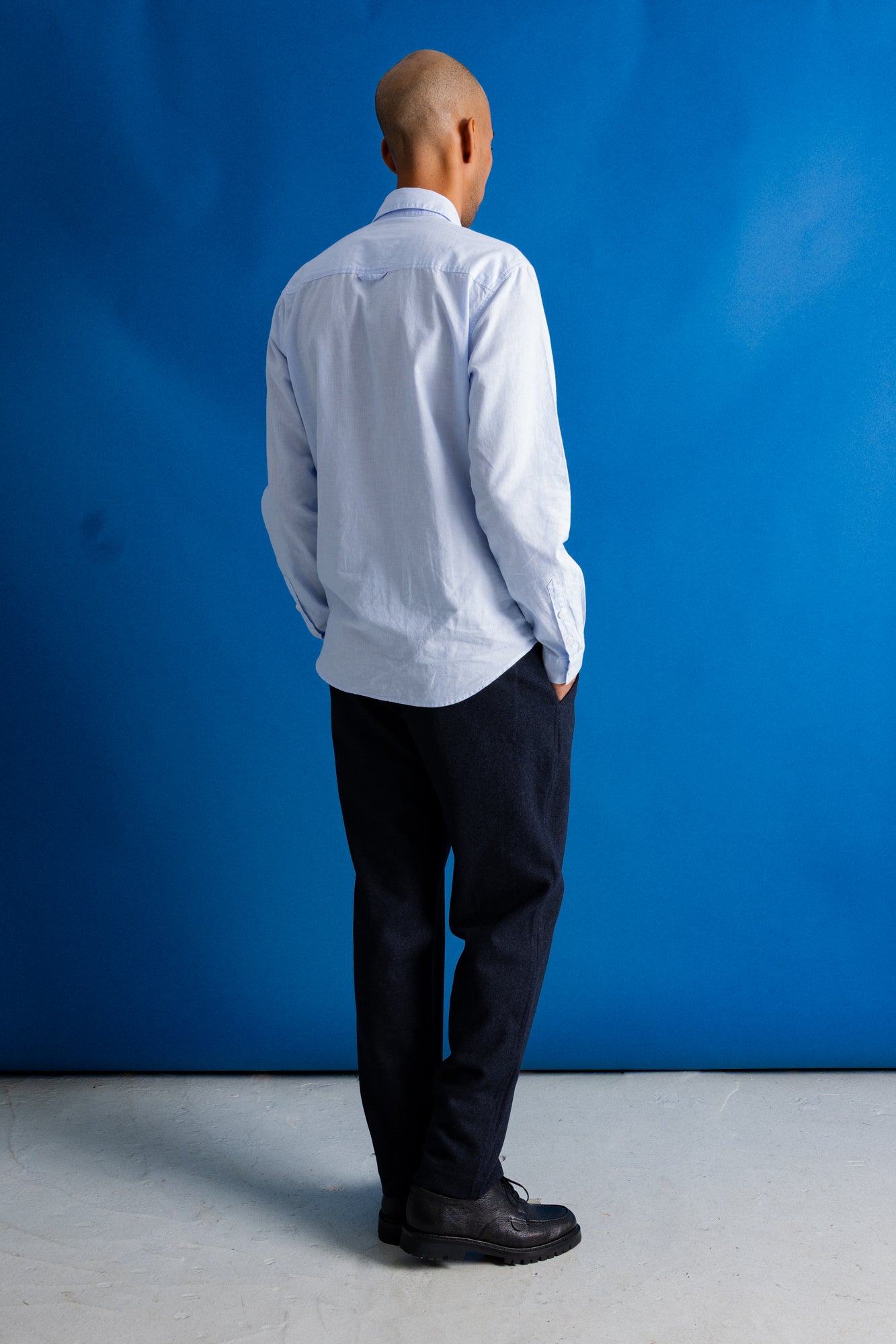 A man viewed from behind wearing a light blue long-sleeve button-down shirt, dark trousers, and black shoes, standing against a bright blue studio backdrop.