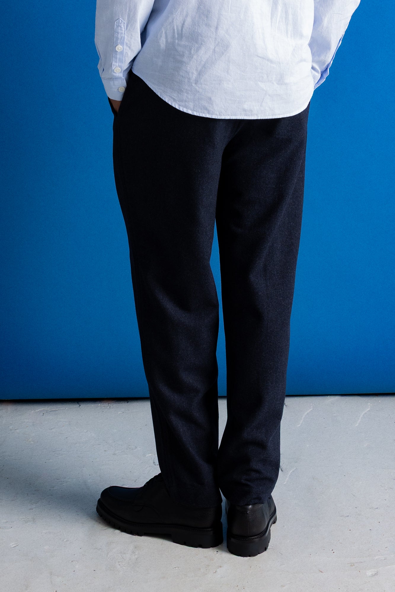 A person shown from the back wearing dark navy trousers, a white shirt, and black shoes, standing against a bright blue wall and light gray floor.