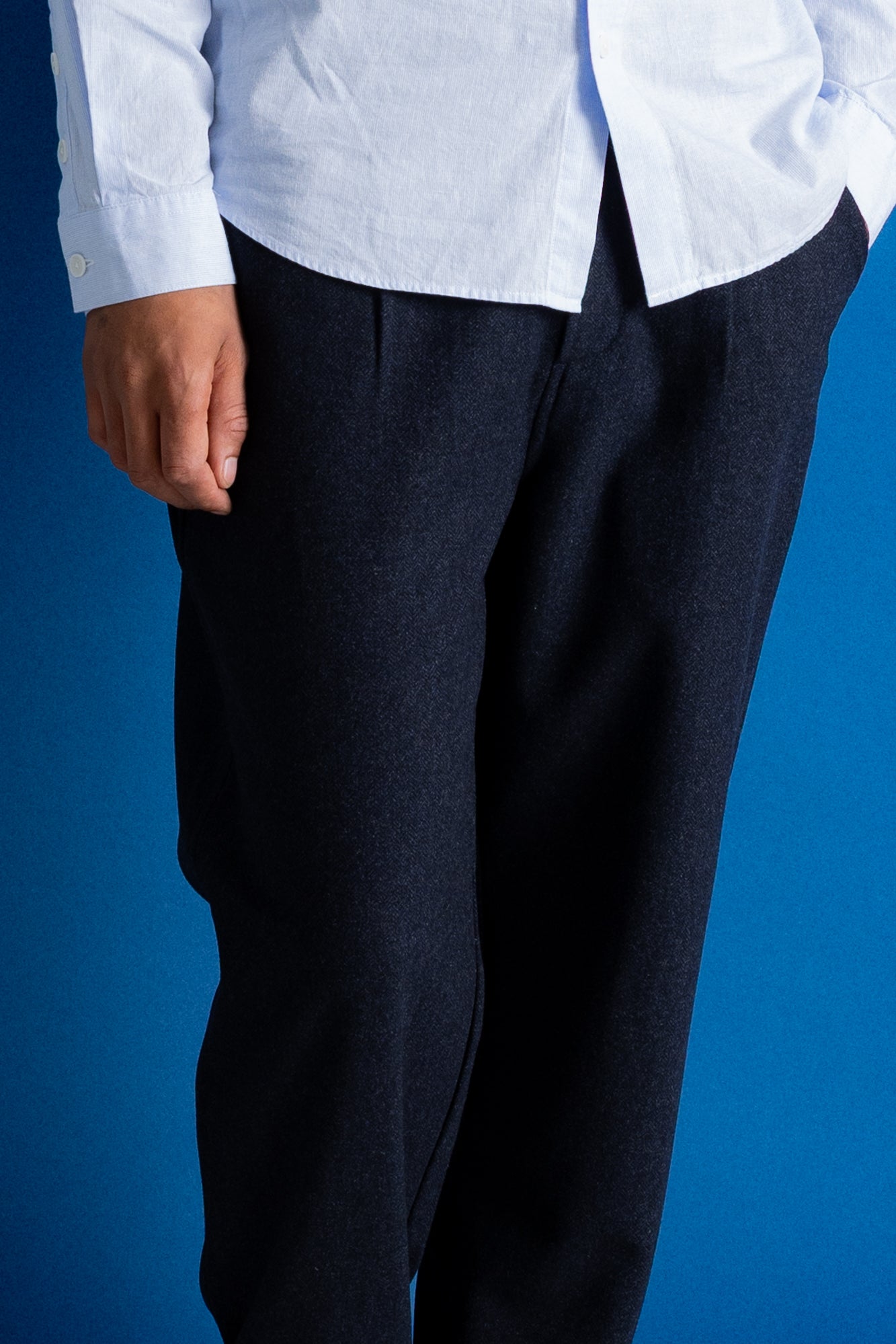 A close-up of a person wearing dark navy trousers and a white button-up shirt against a solid blue background.