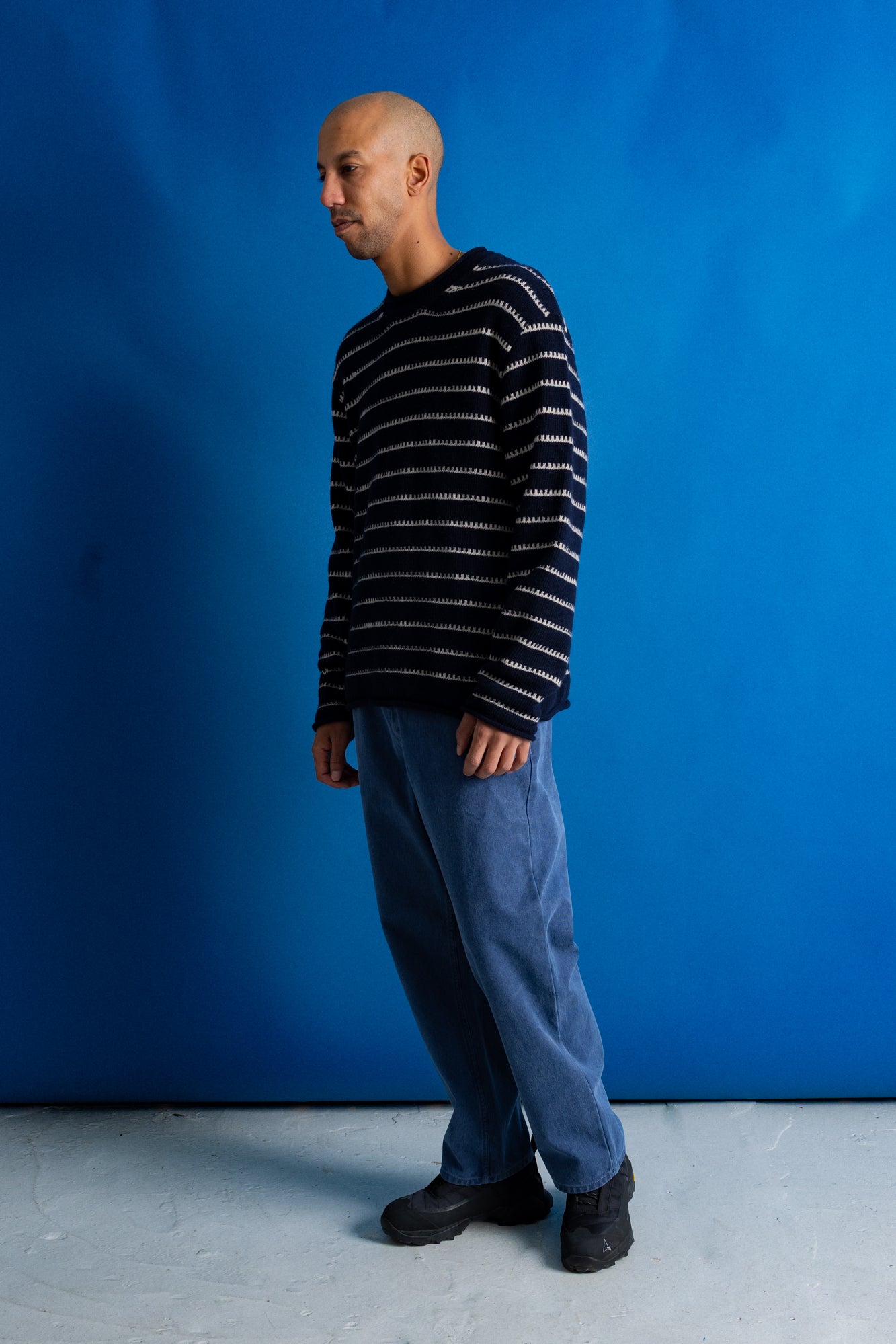 A man wearing a black and white striped knit sweater, blue pants, and black shoes, standing in profile against a solid blue backdrop.