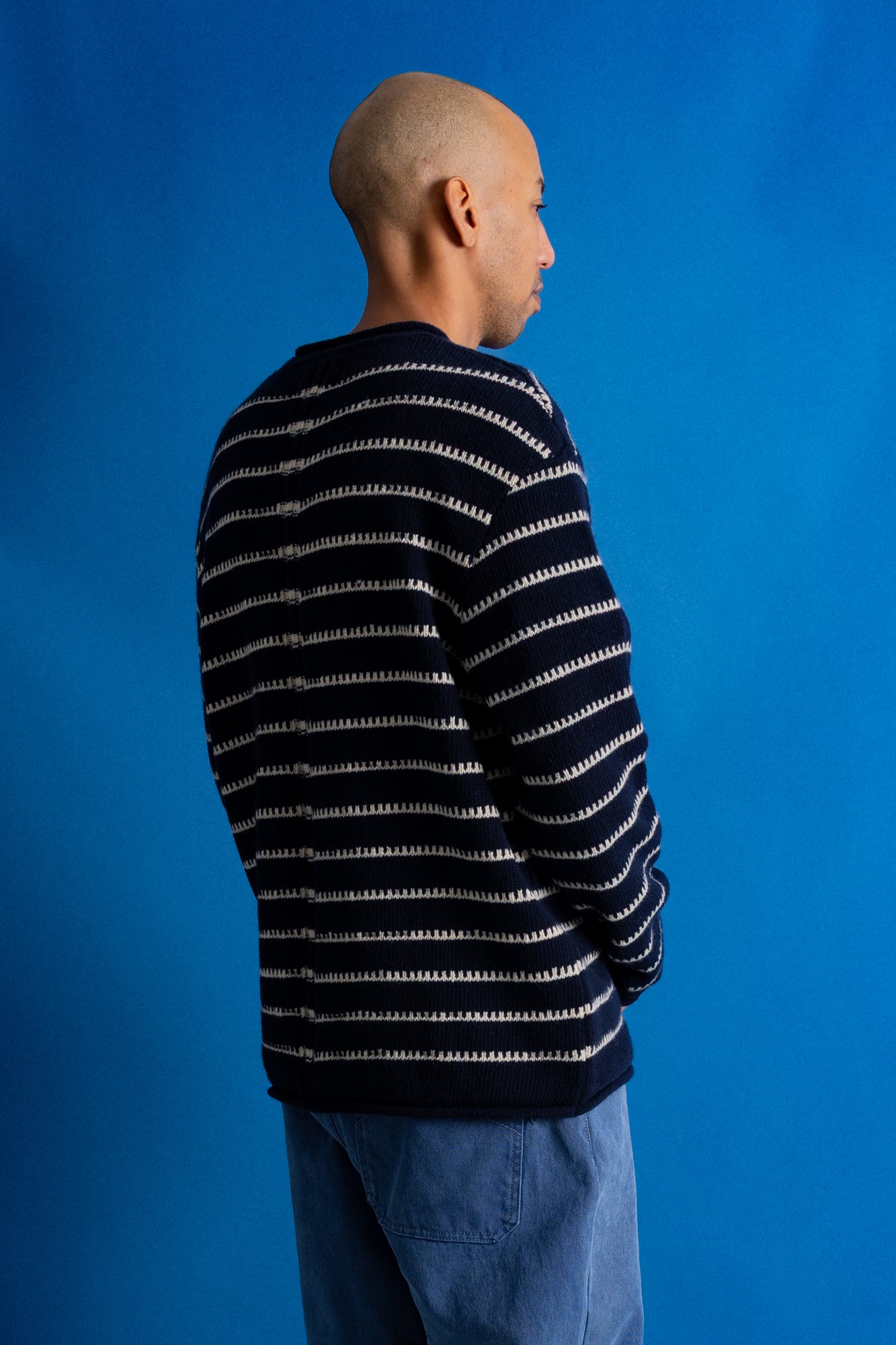 A man wearing a navy blue and white horizontally striped knit sweater, shown from a side-back profile against a solid blue studio background.