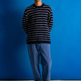 A man wearing a navy and white horizontally striped knit sweater, blue jeans, and black shoes, standing against a solid blue studio backdrop.
