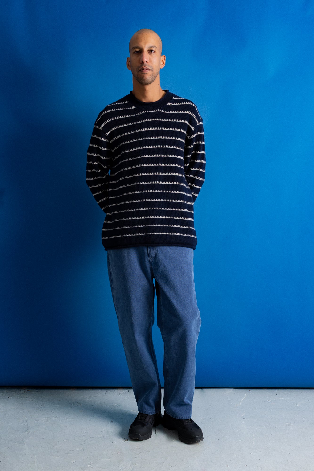 A man wearing a navy and white horizontally striped knit sweater, blue jeans, and black shoes, standing against a solid blue studio backdrop.