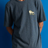 A man wearing a dark heathered blue t-shirt with a small yellow and white lion graphic on the left chest, paired with brown pants, set against a solid blue background.