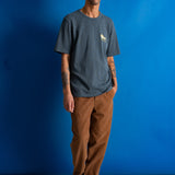 A man stands against a bright blue studio backdrop wearing a dark heather gray crewneck t-shirt with a small yellow graphic on the left chest, brown pants, and black shoes.