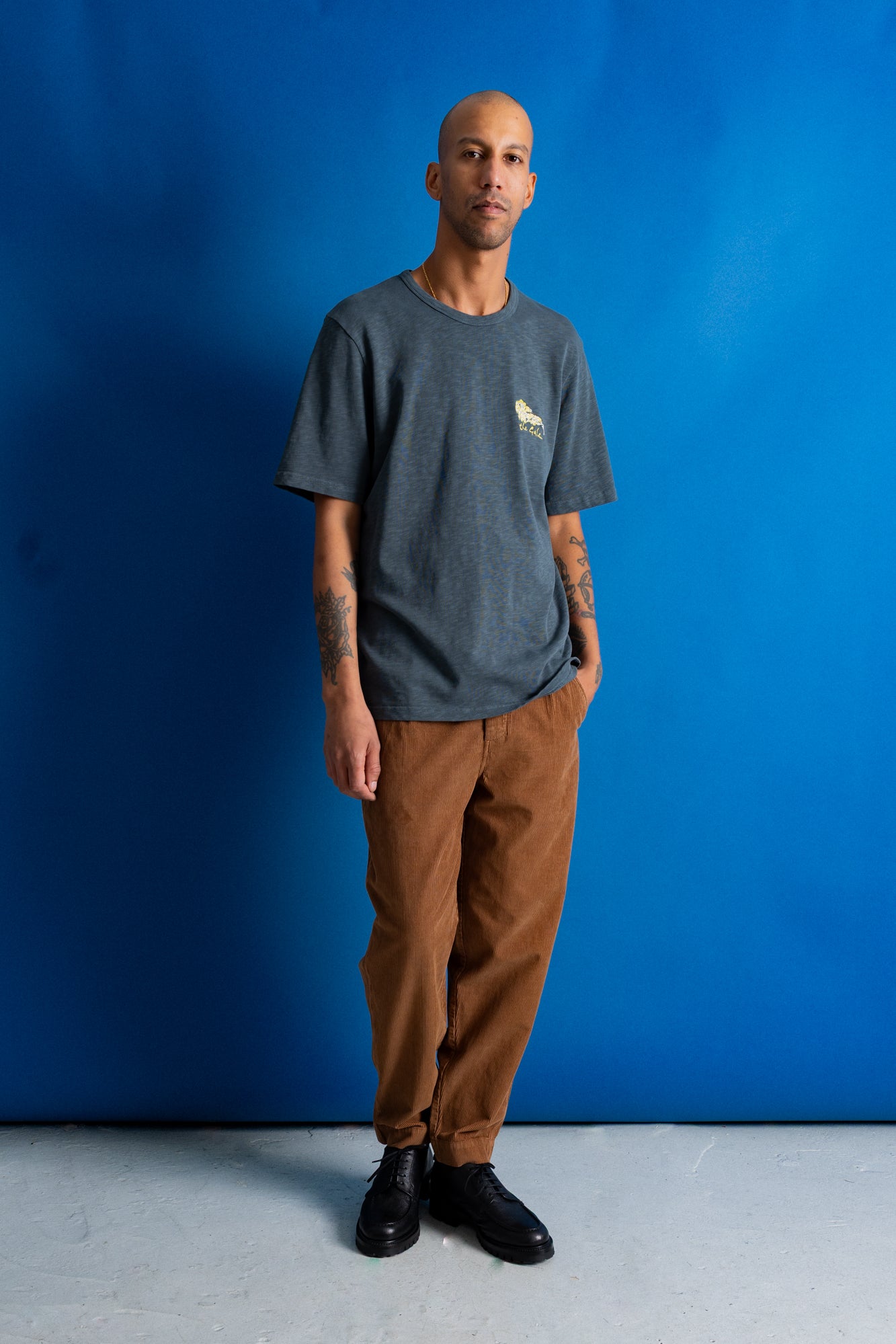 A man stands against a bright blue studio backdrop wearing a dark heather gray crewneck t-shirt with a small yellow graphic on the left chest, brown pants, and black shoes.