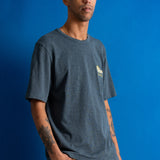 A man stands against a bright blue studio backdrop wearing a dark heather gray crewneck t-shirt with a small yellow graphic on the left chest and orange brown pants.