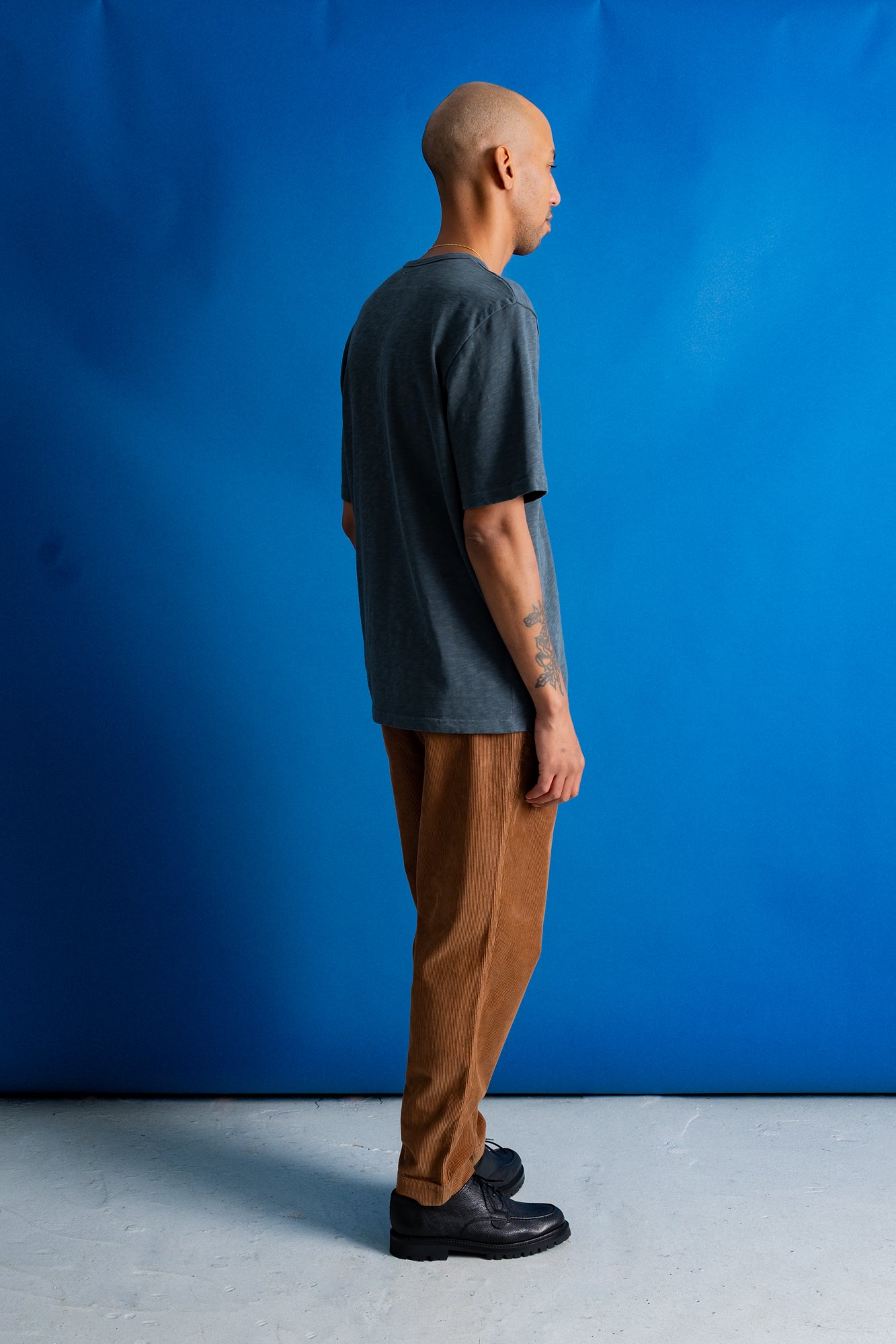 A man stands in profile against a bright blue studio backdrop, wearing a dark charcoal gray t-shirt, brown pants, and black shoes.