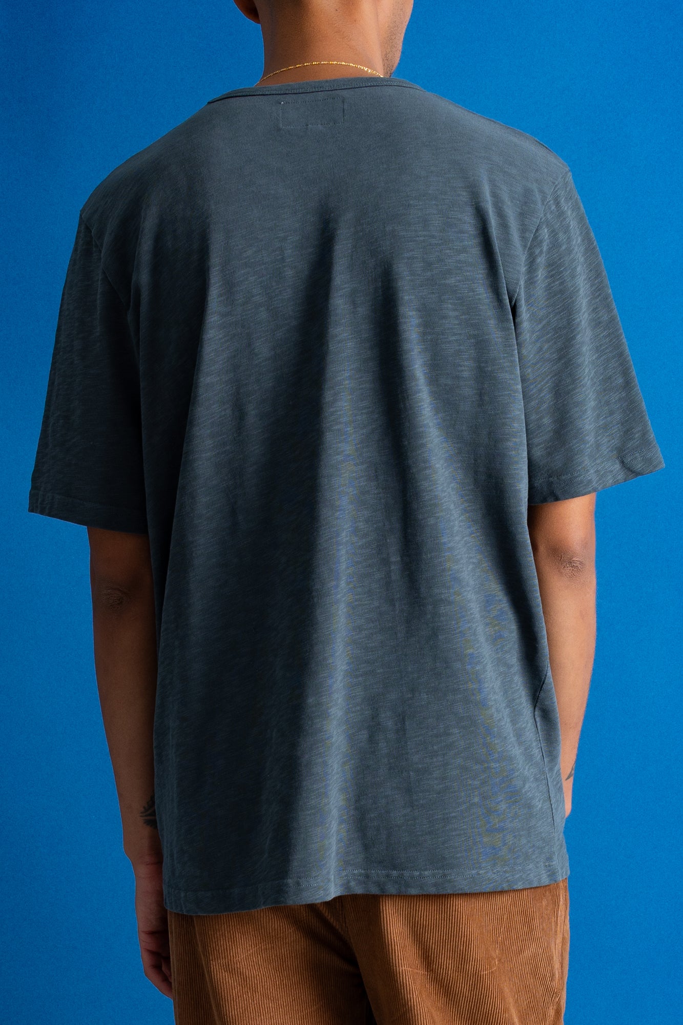 A person shown from the back wearing an oversized slate blue-gray t-shirt and brown pants, set against a solid blue background.