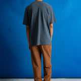 A man viewed from behind wearing a dark gray t-shirt and brown pants, standing against a vibrant blue studio backdrop.