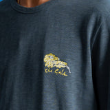 A close-up of a person wearing a dark slate blue heathered t-shirt with a yellow embroidered lion graphic and the text 'the Gale' on the left chest, set against a solid blue background.