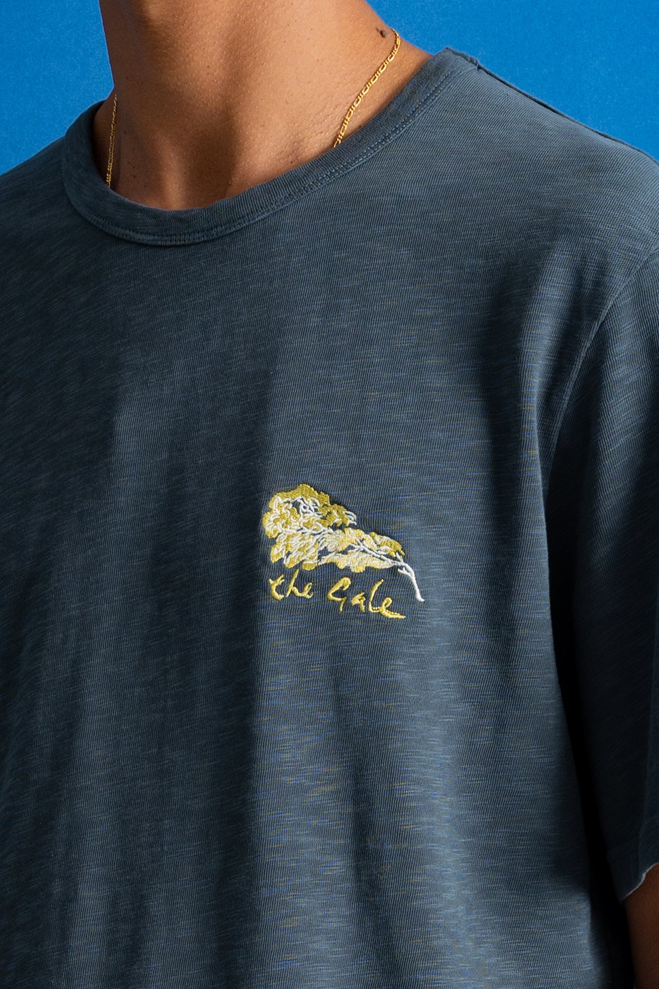 A close-up of a person wearing a dark slate blue heathered t-shirt with a yellow embroidered lion graphic and the text 'the Gale' on the left chest, set against a solid blue background.