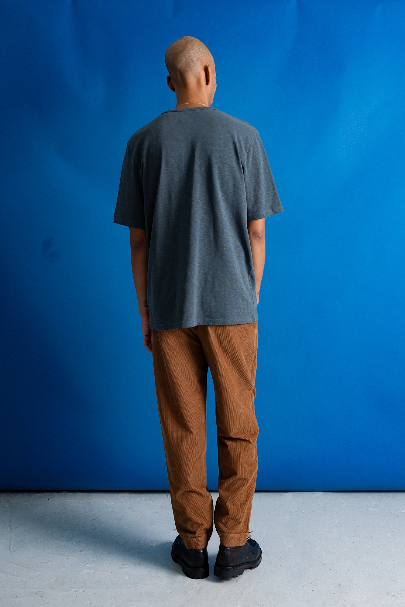 A man stands in profile against a bright blue studio backdrop, wearing a dark charcoal gray t-shirt, brown pants, and black shoes.