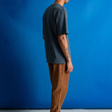 A man stands in profile against a bright blue studio backdrop, wearing a dark charcoal gray t-shirt, brown pants, and black shoes.