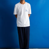 A man stands against a bright blue studio backdrop wearing a white short-sleeve crewneck T-shirt with a small light blue graphic on the left chest, paired with dark navy pants and black shoes.