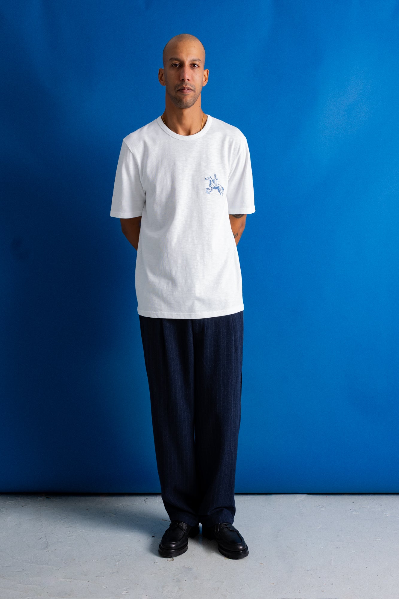 A man stands against a bright blue studio backdrop wearing a white short-sleeve crewneck T-shirt with a small light blue graphic on the left chest, paired with dark navy pants and black shoes.