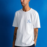 A man wearing a white crewneck t-shirt with a small blue graphic on the left chest, paired with dark blue pants, standing against a solid blue background.