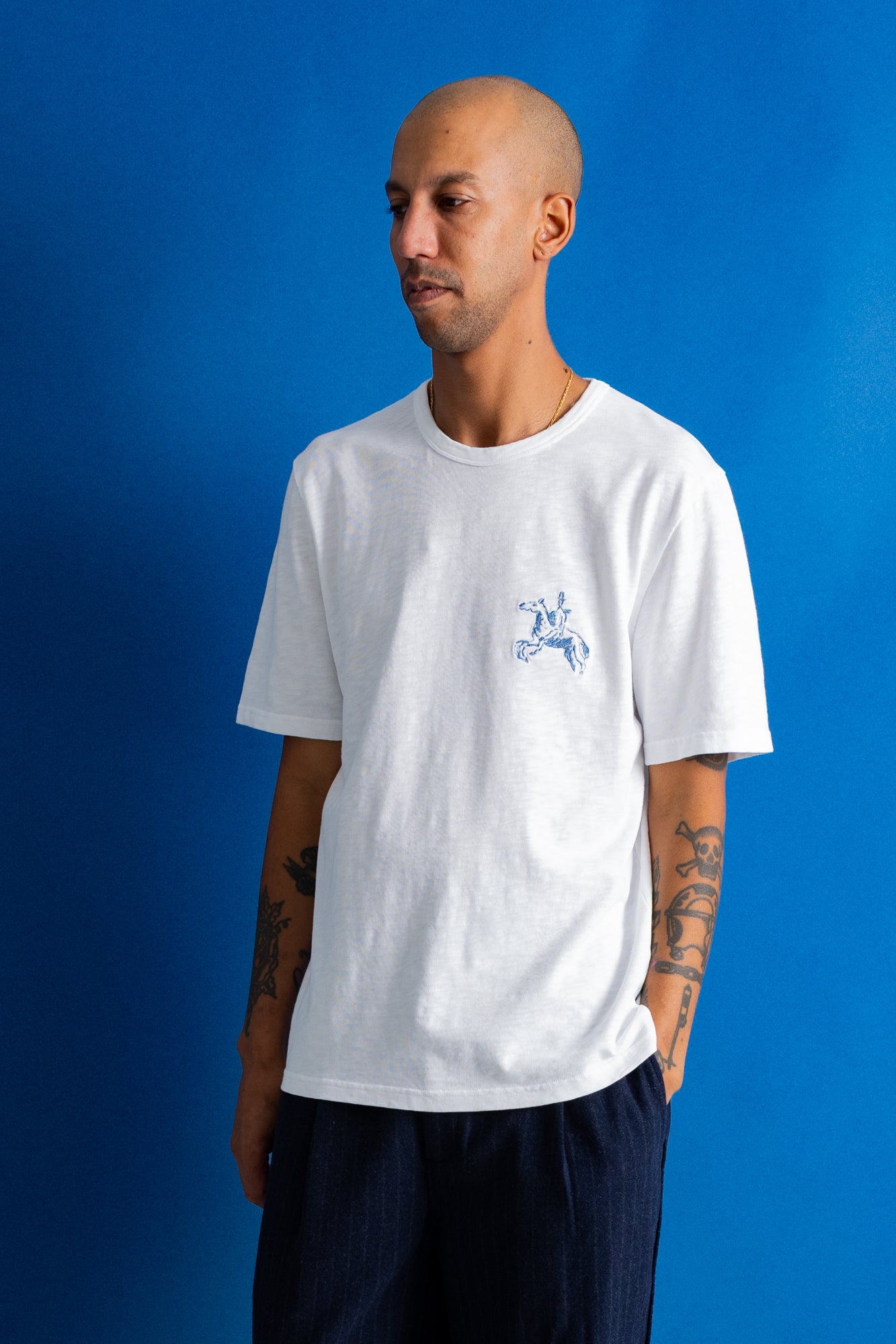 A man wearing a white crewneck t-shirt with a small blue graphic on the left chest, paired with dark blue pants, standing against a solid blue background.