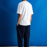 A man viewed from behind wearing a white short-sleeve t-shirt and dark navy blue relaxed-fit pants, standing against a solid blue studio backdrop.