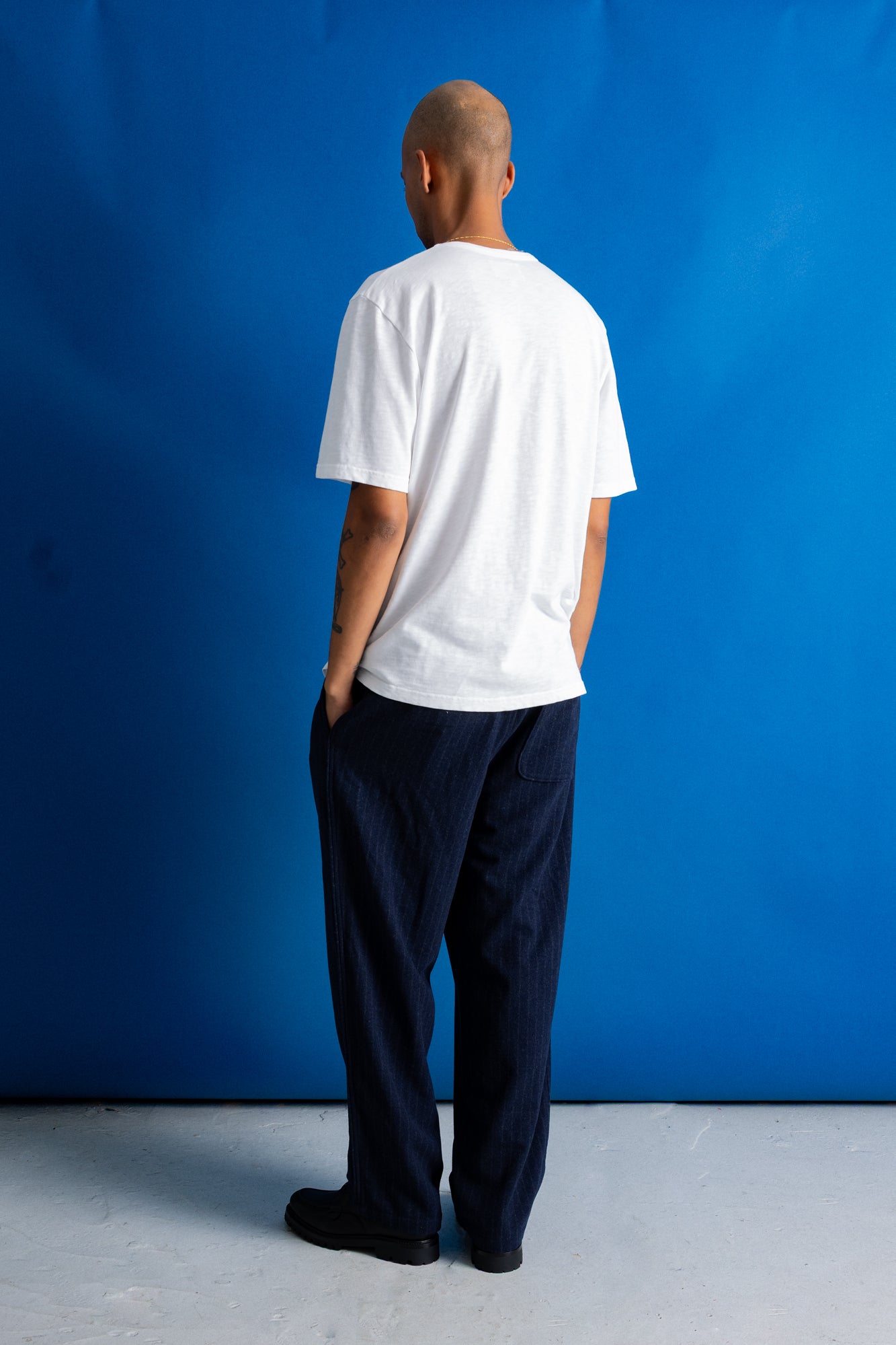 A man viewed from behind wearing a white short-sleeve t-shirt and dark navy blue relaxed-fit pants, standing against a solid blue studio backdrop.
