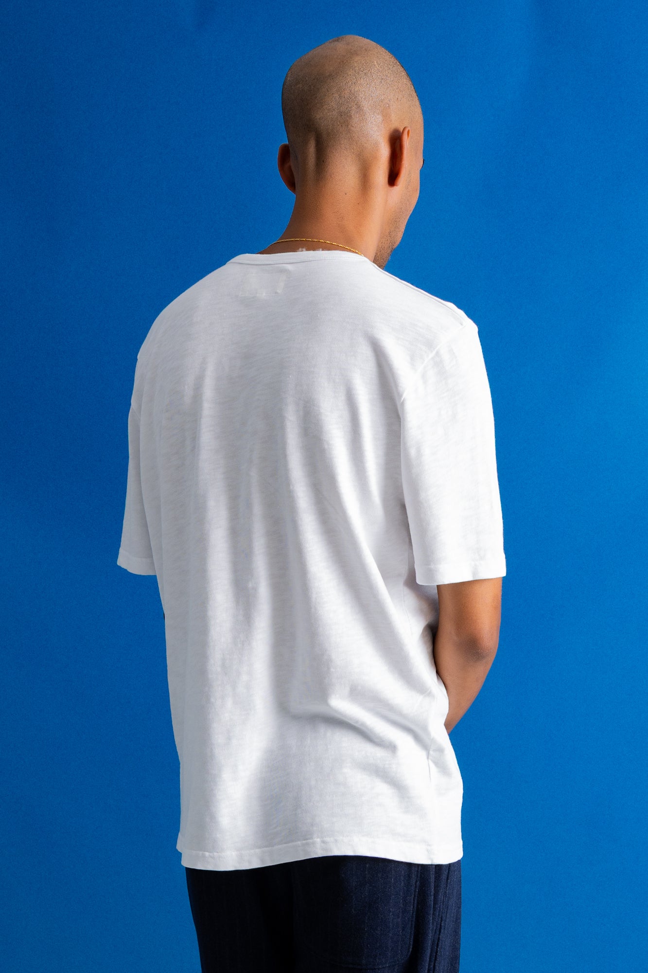 A man viewed from behind wearing a plain white short-sleeve t-shirt and dark pants, standing against a solid blue background.