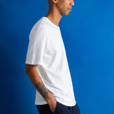 A man in profile wearing a white short-sleeve crewneck t-shirt and dark navy pants, standing against a solid blue studio background.