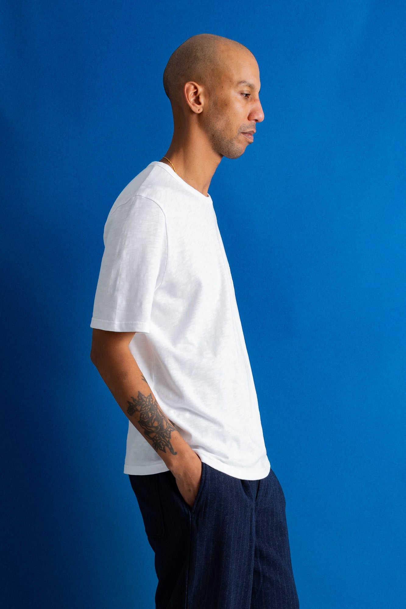 A man in profile wearing a white short-sleeve crewneck t-shirt and dark navy pants, standing against a solid blue studio background.