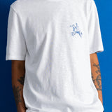 A man wearing a white crewneck t-shirt with a small blue embroidered cowboy on a horse logo on the left chest, set against a solid blue background.