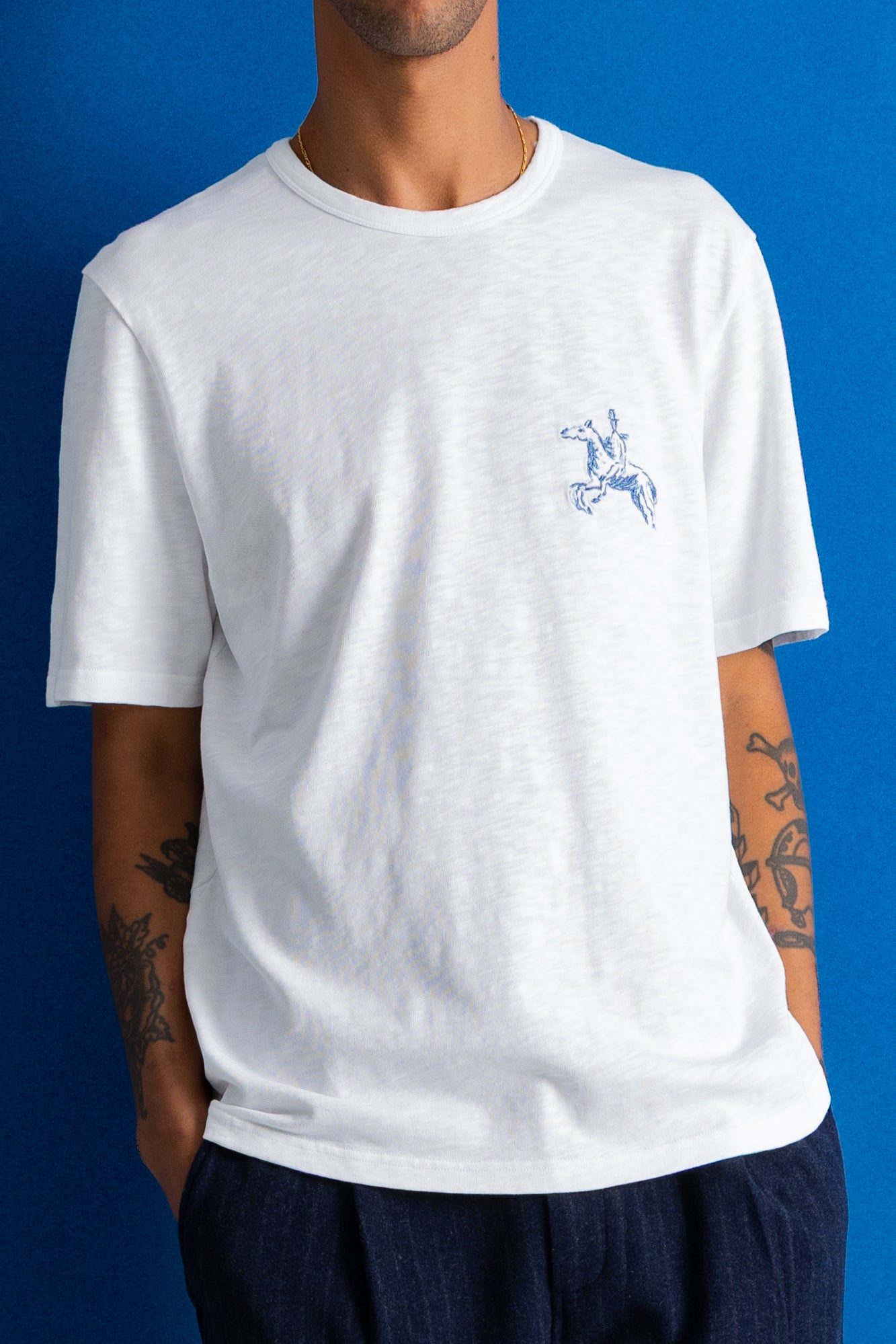A man wearing a white crewneck t-shirt with a small blue embroidered cowboy on a horse logo on the left chest, set against a solid blue background.