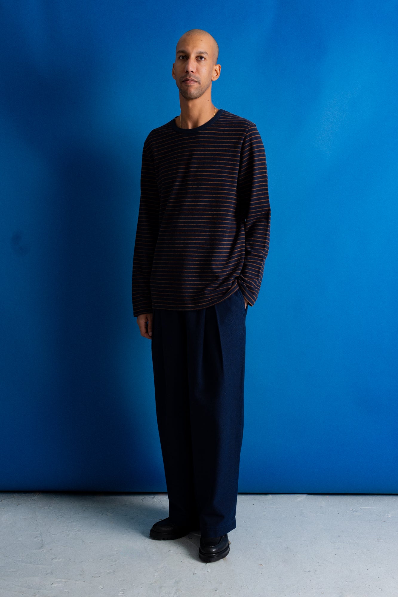 A man stands against a bright blue studio backdrop wearing a dark navy long-sleeve shirt with thin red horizontal stripes and dark navy wide-leg trousers.