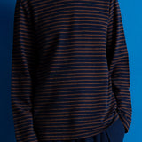 A person wearing a dark navy long-sleeve knit top with thin horizontal rust-orange stripes, paired with dark navy trousers, set against a solid blue background.