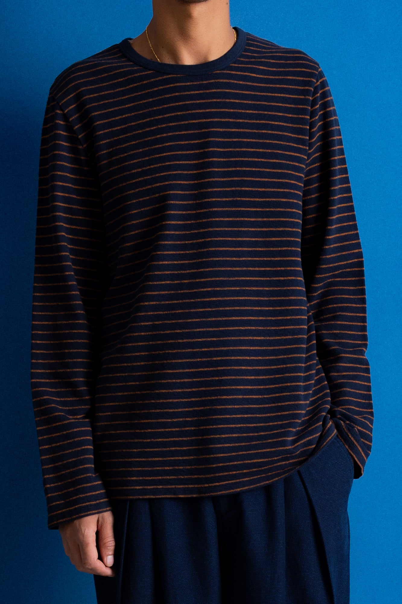A person wearing a dark navy long-sleeve knit top with thin horizontal rust-orange stripes, paired with dark navy trousers, set against a solid blue background.