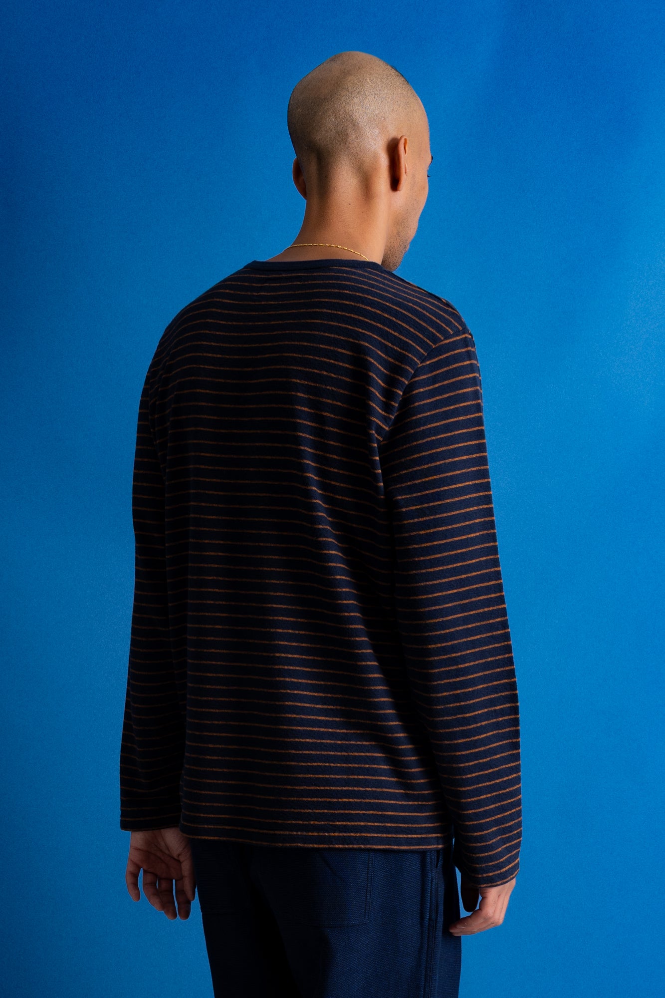 A man viewed from behind wearing a dark navy long-sleeve shirt with thin orange horizontal stripes, set against a solid blue background.