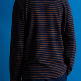 A person shown from the back wearing a dark navy long-sleeve shirt with thin orange horizontal stripes, paired with dark blue pants, against a solid blue studio background.