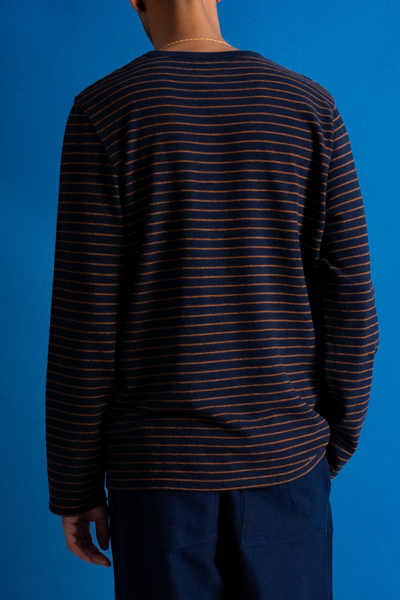 A person shown from the back wearing a dark navy long-sleeve shirt with thin orange horizontal stripes, paired with dark blue pants, against a solid blue studio background.