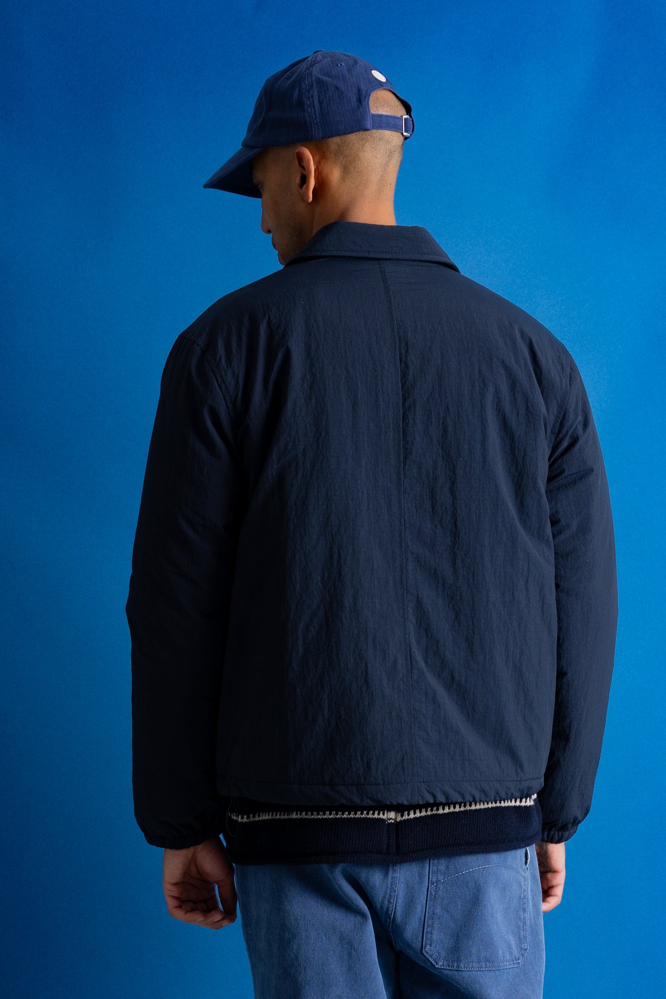 A man viewed from behind wearing a dark navy blue jacket, blue jeans, and a navy blue baseball cap, set against a solid blue studio background.