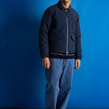 A man stands against a bright blue studio backdrop wearing a dark navy zip-front jacket with a hood, blue jeans, black shoes, and a navy cap.